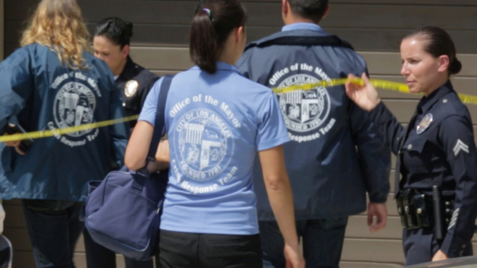 CRT Volunteers wearing blue shirts cross a yellow crime tape held by an LAPD officer